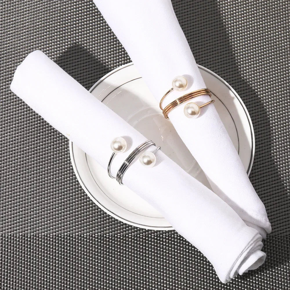 White cloth napkins rolled with pearl-accented gold and silver napkin rings on a white plate