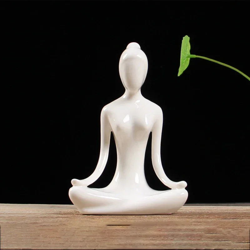 White ceramic sculpture of a person in lotus yoga pose on wooden surface with black background and green leaf