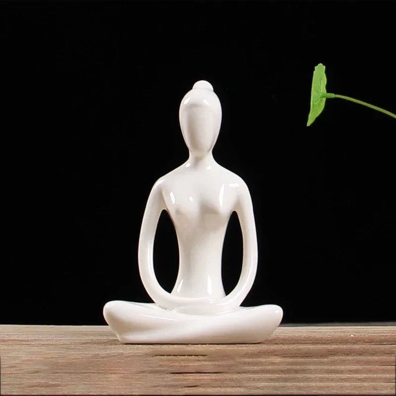 Minimalist white ceramic sculpture of a meditating figure with a green leaf on wooden surface