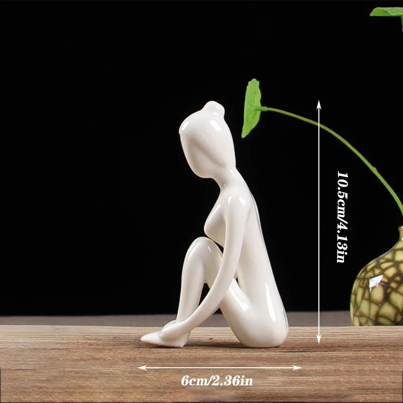 Glossy white ceramic figurine of a seated abstract human figure, measuring 10.5cm tall and 6cm wide, on wooden surface