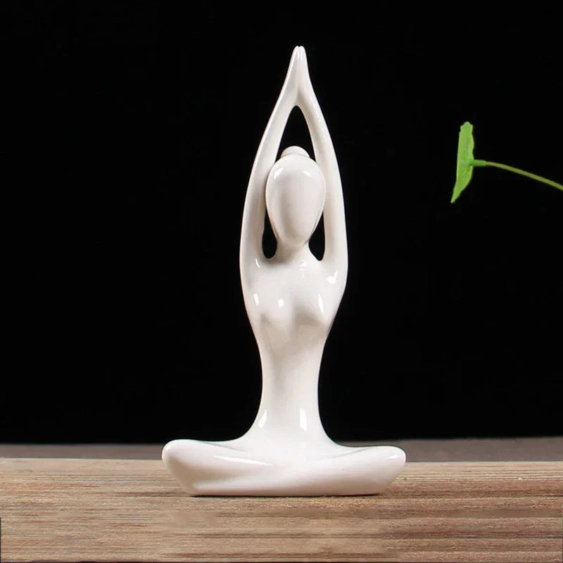 Minimalist white ceramic yoga figure sculpture sitting cross-legged with hands raised, on wooden surface